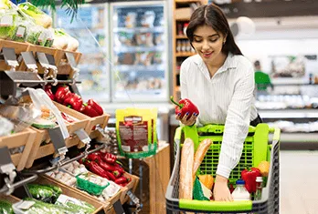 A woman shopping in a grocery store aisle filled with fresh vegetables, placing a red bell pepper into a green shopping cart that already contains baguettes, milk, and other groceries.
