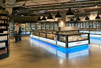 Modern grocery store interior featuring refrigerated display cases lit with blue LED lighting, stocked with various food items, and a customer browsing products in the background.