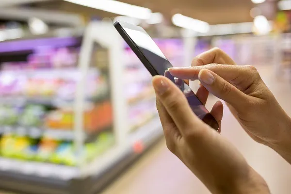 Person using a smartphone with one hand tapping the screen in a grocery store aisle with blurred shelves behind.