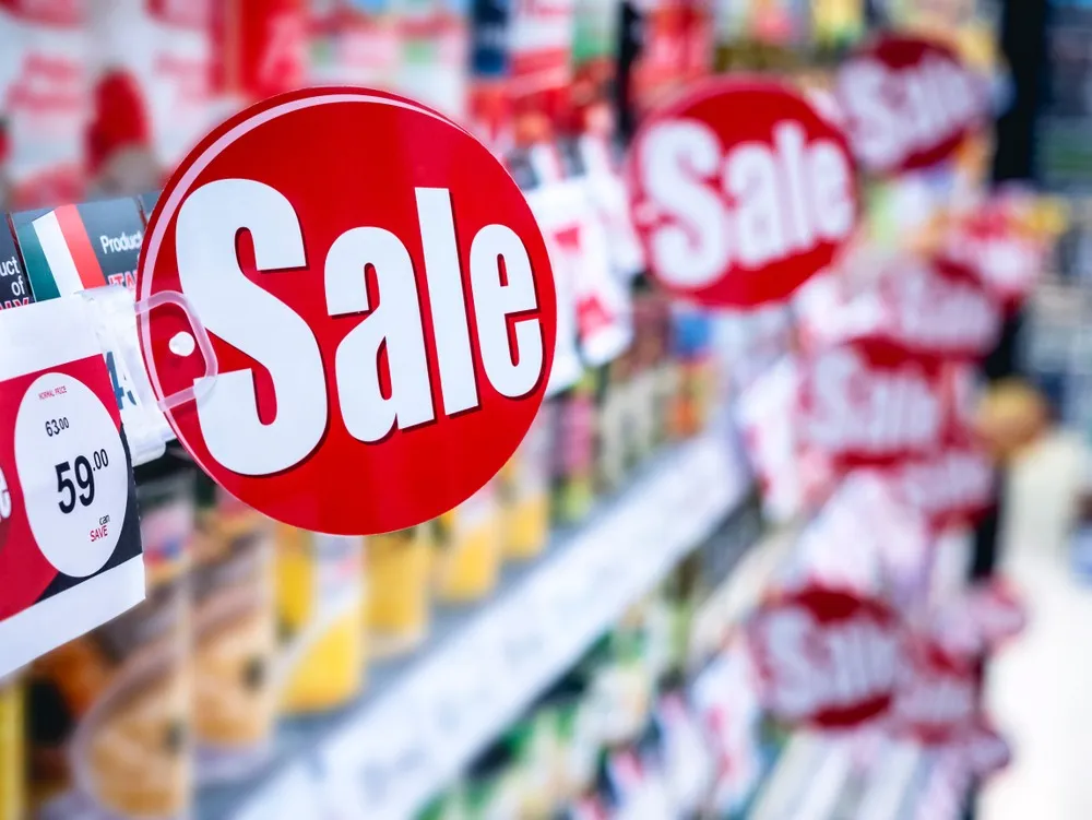 Close-up of a red circular sale sign with white text on a store shelf, showing discounted price tags and blurred products in the background.