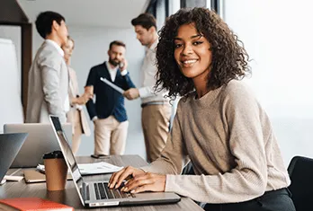 Smiling woman with curly hair sitting at a desk using a laptop, with three colleagues standing and talking in the background of a modern office.