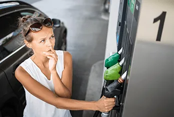 A woman with sunglasses on her head stands by a gas pump at a station, holding the fuel nozzle and looking thoughtfully at the pump display.