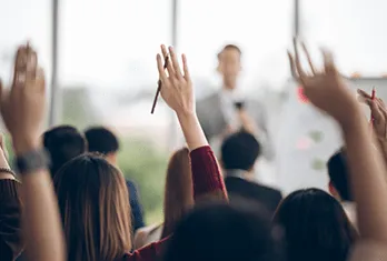 Audience raising hands in a classroom or seminar setting with a speaker blurred in the background.