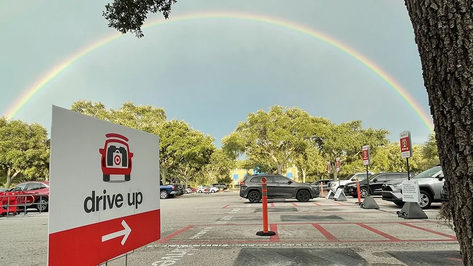 Parking lot with a drive-up sign featuring a red car icon with a camera, pointing right, under a clear sky with a bright full rainbow arching over trees and parked cars.