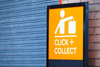 Orange and black sign with a white icon of a person holding a shopping bag and text reading 'CLICK + COLLECT' on a background of a blue shutter door and brick wall.