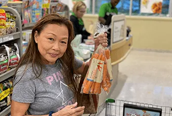 Woman holding a bag of carrots inside a grocery store near a shopping cart filled with items.