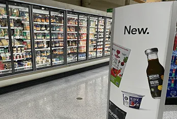 Supermarket aisle with refrigerated glass doors containing various packaged food products and a white display stand in the foreground featuring the word 'New' and images of a beverage bottle and yogurt containers.