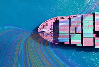 Aerial view of a cargo ship loaded with colorful shipping containers moving through bright blue water with multi-colored trails behind the ship.