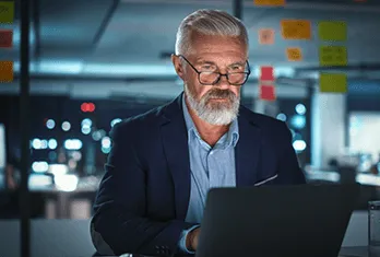 Older man with gray hair and beard wearing glasses and a navy blazer working on a laptop in a dimly lit office with sticky notes on glass wall behind him.
