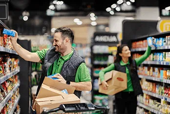 Smiling grocery store employees wearing green uniforms stocking shelves with products in aisles of a store.
