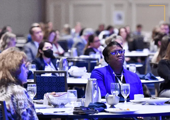 Audience of diverse adults seated at round tables attending a conference or seminar in a large room, listening attentively.