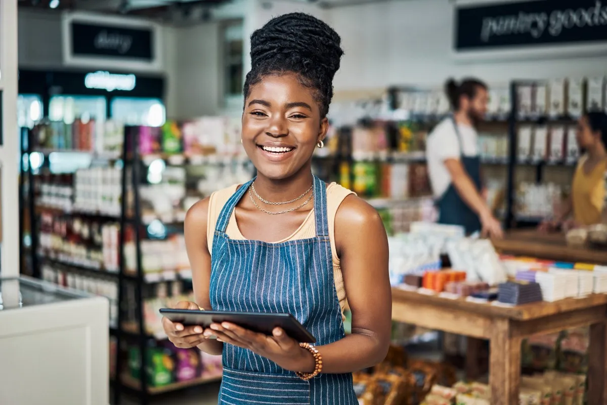 Smiling young woman wearing a striped apron holding a tablet in a grocery store with shelves and products in the background.