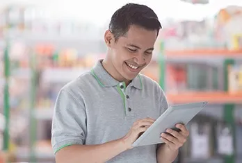 Smiling man using a tablet device in a brightly lit store with shelves of products in the background.