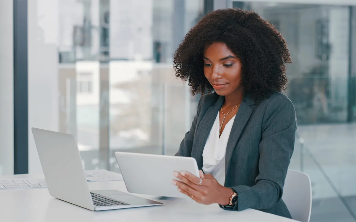 Professional woman with curly hair in a gray blazer sitting at a white desk in a modern office, looking at a tablet with a laptop beside her.