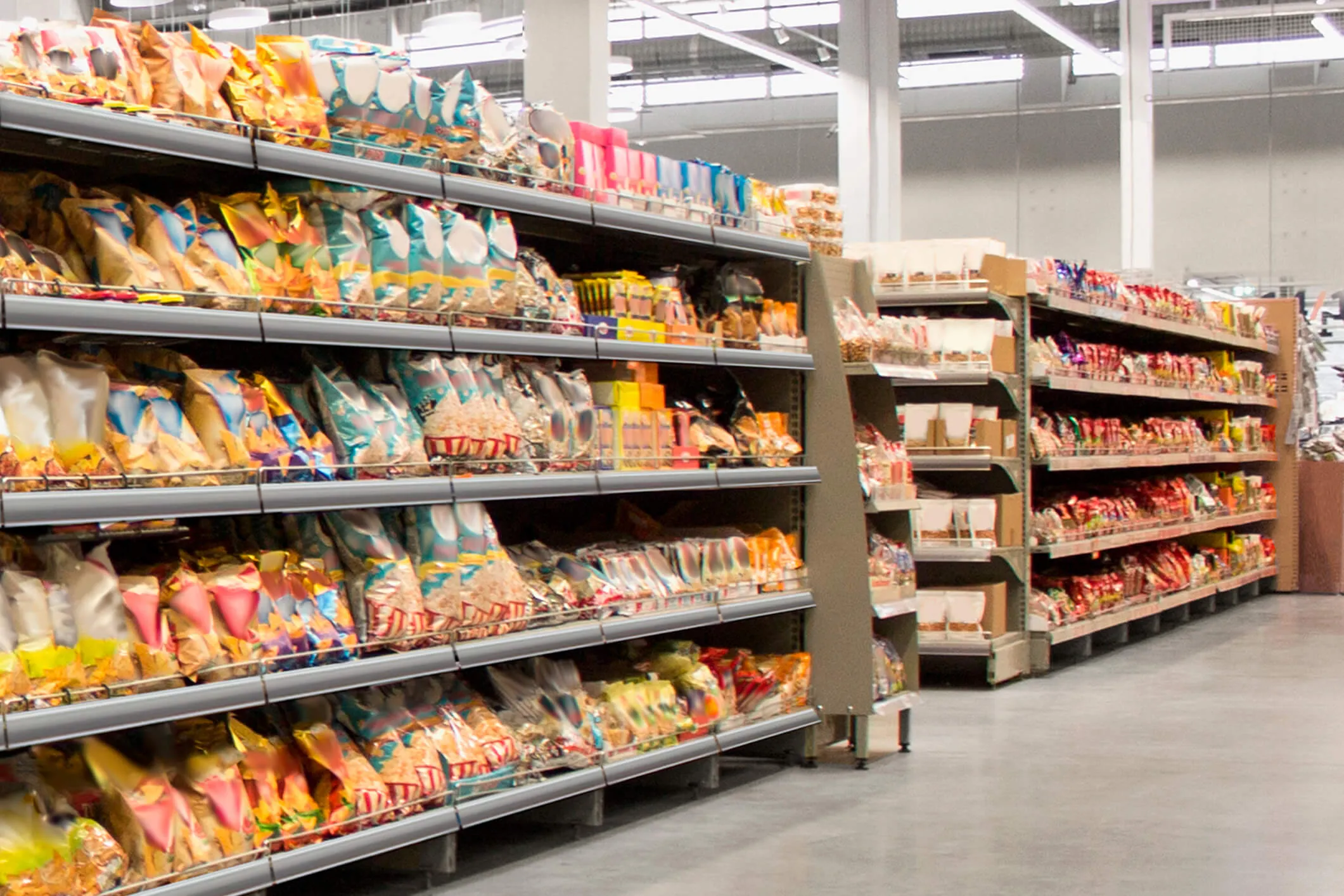 Aisle in a warehouse-style store with metal shelves stocked with various colorful packaged snacks and food items, under bright industrial lighting.
