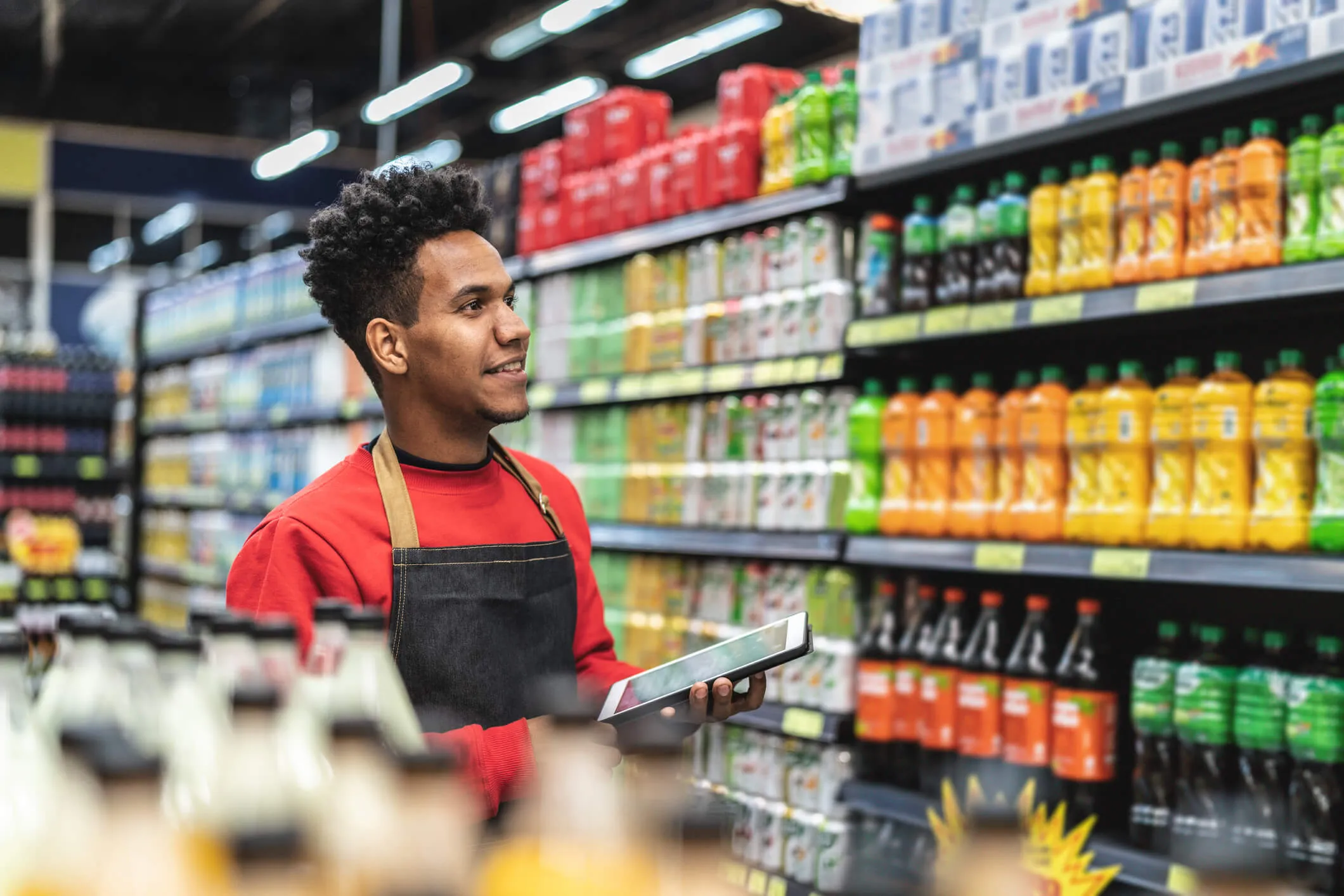 A smiling male grocery store employee wearing a red shirt and black apron holds a tablet while standing in an aisle stocked with colorful bottled beverages.