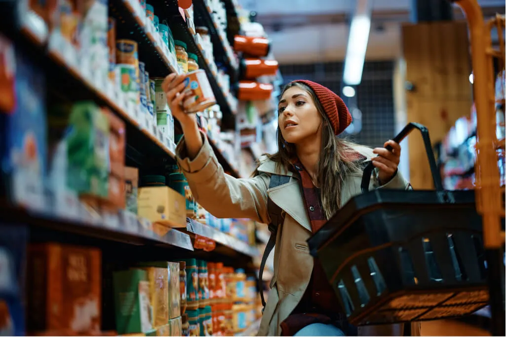A woman wearing a red beanie and beige trench coat shops in a grocery store aisle, holding a black basket and reaching for a canned product on a shelf filled with various food items.