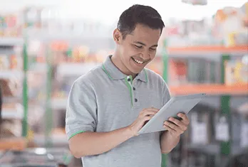 Man in a gray polo shirt smiling while using a tablet in a brightly lit store interior with shelves.