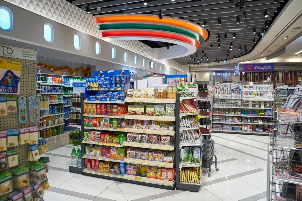 Interior of a brightly lit convenience store with shelves stocked with snacks, beverages, magazines, and other products under a ceiling with colorful circular lighting and small window-like fixtures.