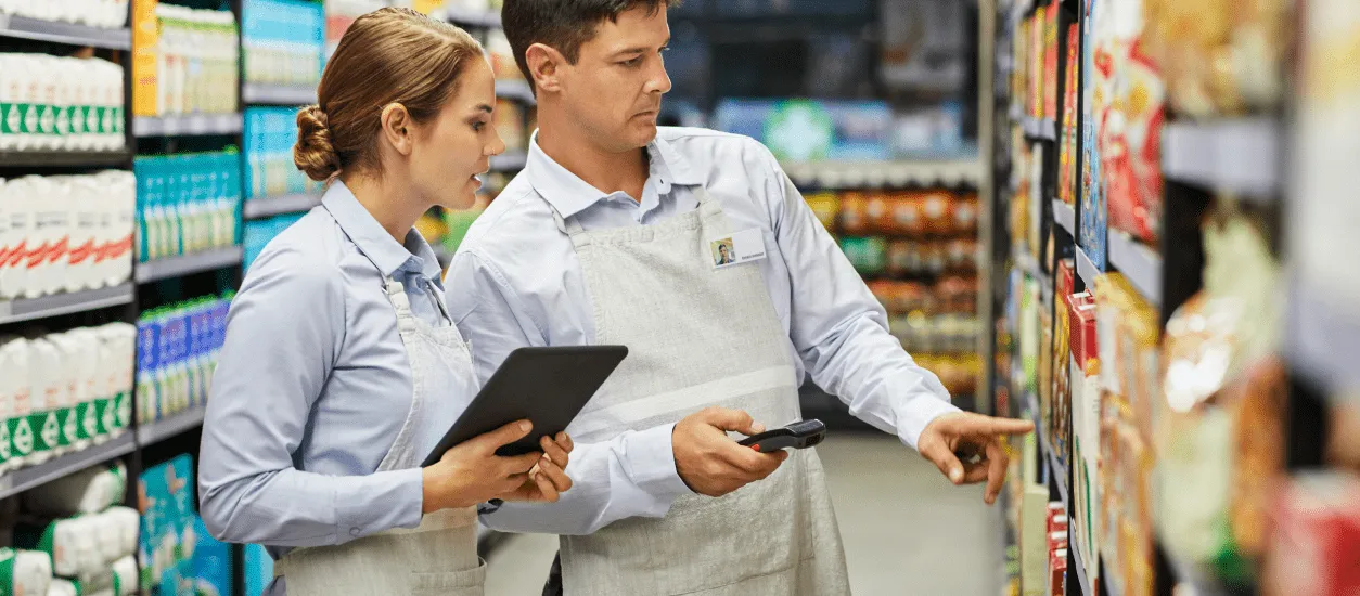 Two grocery store workers wearing aprons and light blue shirts stand in an aisle filled with packaged goods; the man points to a product on the shelf while holding a scanner, and the woman holds a tablet, both seemingly reviewing inventory.