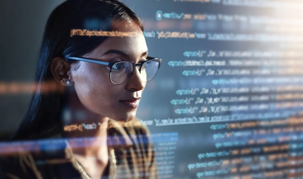 Woman with glasses focused on computer screen displaying lines of code in a dimly lit environment.