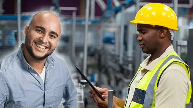 Two male factory workers in a manufacturing setting: one smiling and facing the camera while the other wearing a yellow hard hat and safety vest uses a digital tablet.