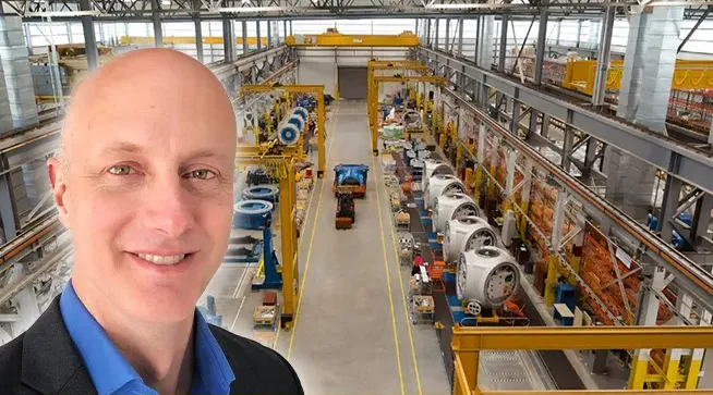 Smiling bald man in a blue shirt and dark blazer superimposed over a large industrial warehouse with yellow machinery and equipment arranged along a central aisle.