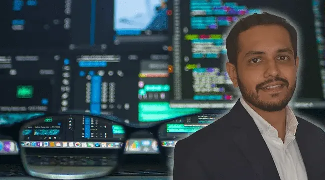 Man with short dark hair and beard wearing a black suit jacket and white shirt, smiling in front of a blurred background of computer screens with colorful code and data displays, with the foreground showing a blurred pair of eyeglasses focusing on a computer screen displaying data and code.