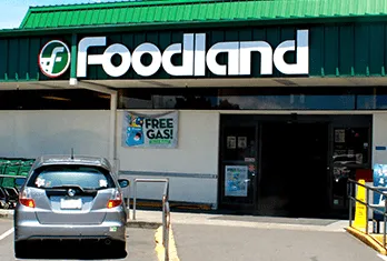 Exterior of a Foodland grocery store with a green roof and a large Foodland sign above the entrance, a silver car parked in front, and a sign on the wall advertising free gas.