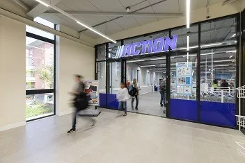 Entrance of an Action store with glass doors, a large blue and white sign above, and people walking in and out, some blurred in motion.