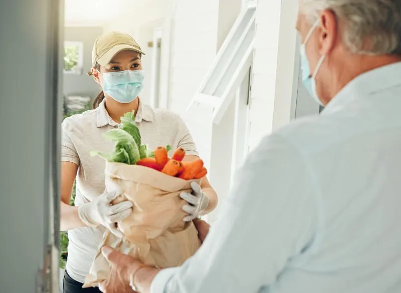 Masked delivery woman wearing a cap and gloves handing a paper bag full of fresh vegetables like carrots and lettuce to an elderly man wearing a mask at his doorway.
