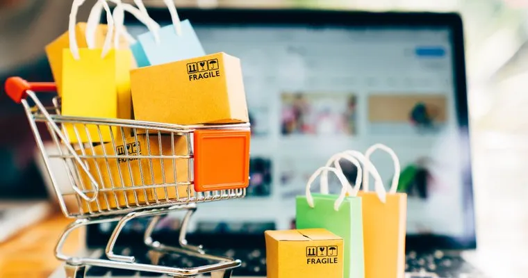 Small shopping cart filled with colorful shopping bags and cardboard boxes labeled fragile, placed on a laptop keyboard with a blurred website on the laptop screen.