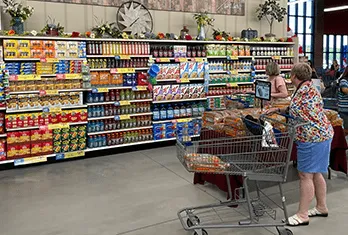 Senior woman with a shopping cart stands in front of well-stocked supermarket shelves filled with colorful packaged food products, including cereals, snacks, and canned goods. Another shopper is visible nearby.