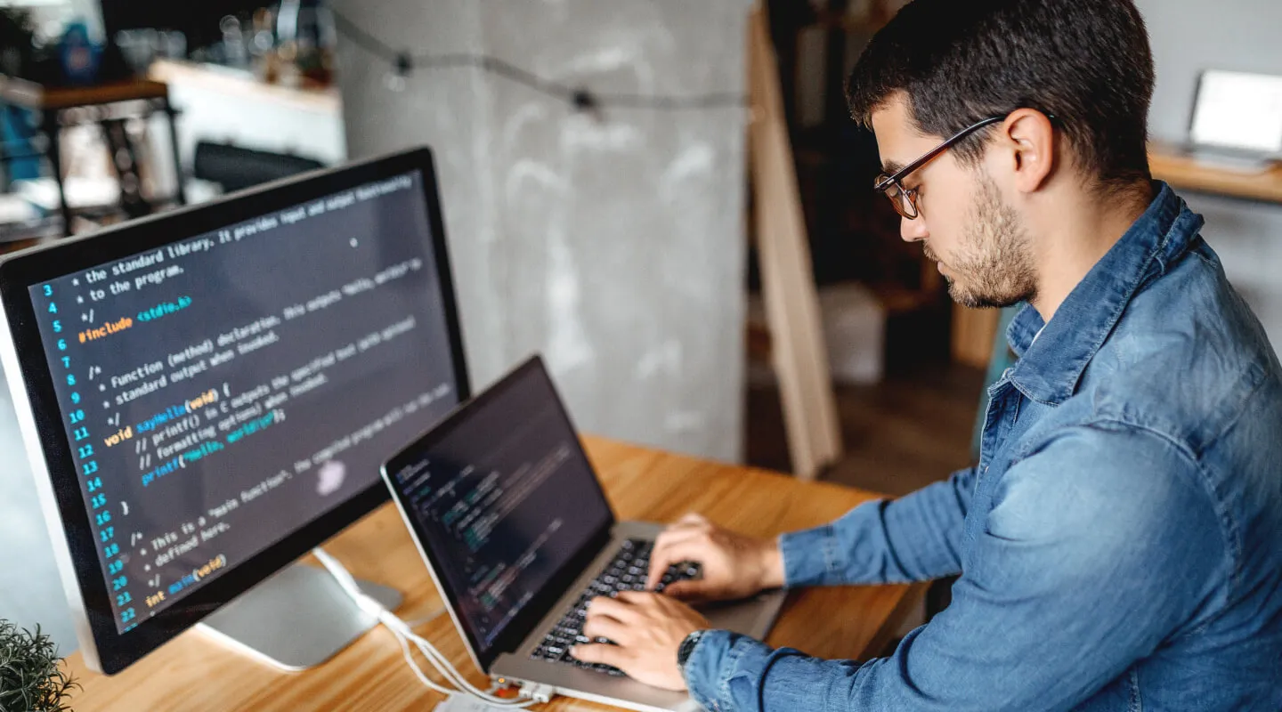 Man wearing glasses and a blue denim shirt typing on a laptop at a wooden desk with a large monitor displaying C programming code.