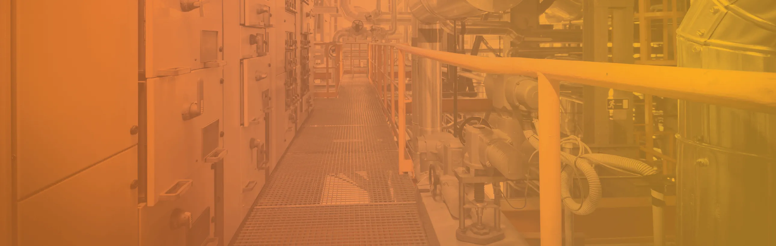 Industrial walkway with metal grating floor and yellow handrails, surrounded by pipes, valves, and control panels in a factory setting.