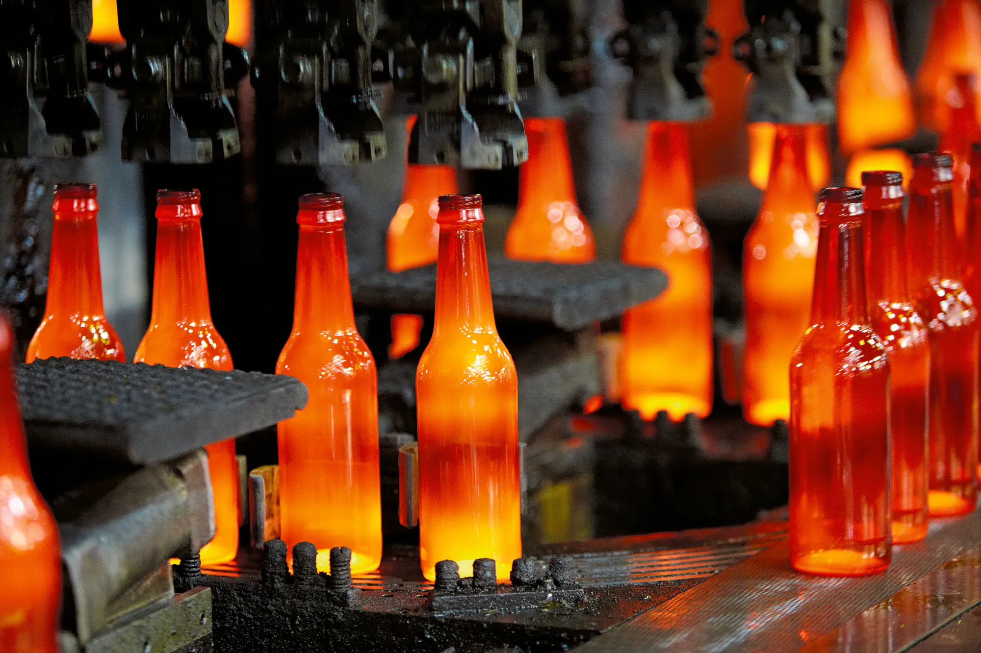 Rows of glowing orange molten glass bottles on a conveyor belt in a glass manufacturing furnace.