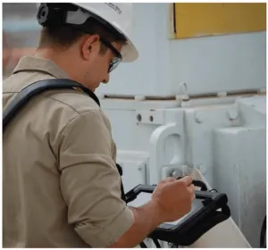 Worker wearing a white helmet and safety glasses using a tablet device in an industrial setting with machinery.