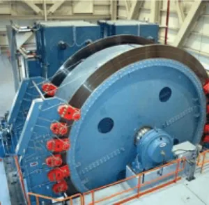 Large industrial machine with two massive blue circular wheels mounted horizontally and connected, surrounded by red components and railings in a factory setting.