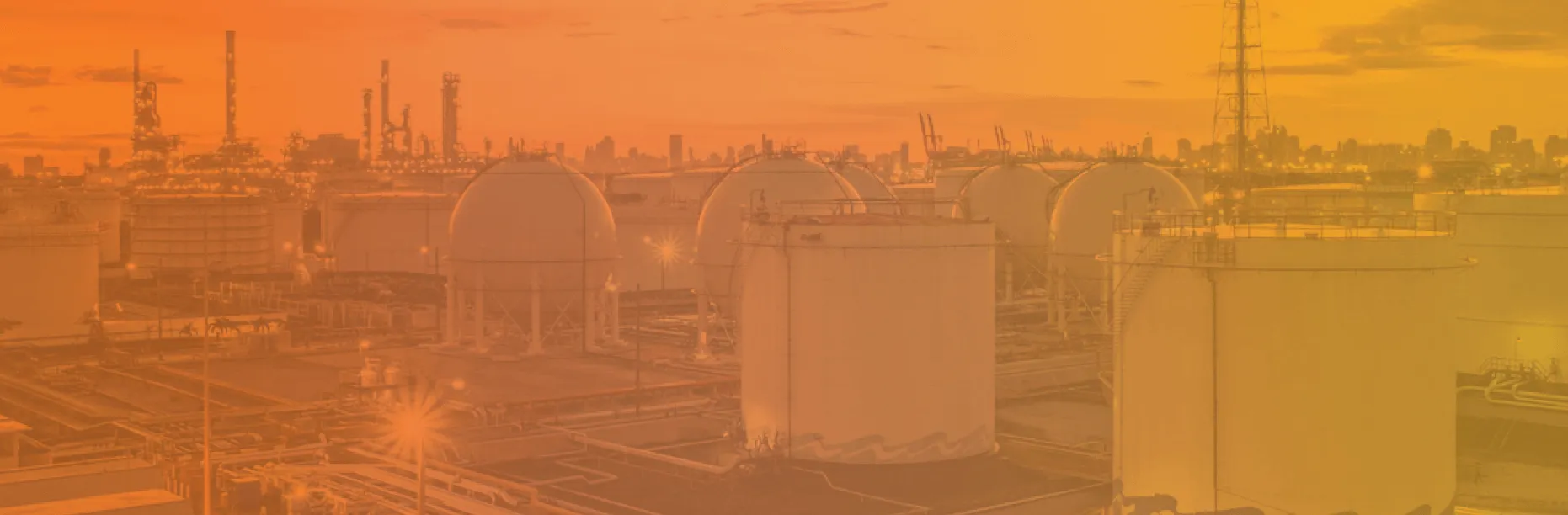 Oil refinery and storage tanks at dusk with industrial structures and city skyline in the background under an orange sky.