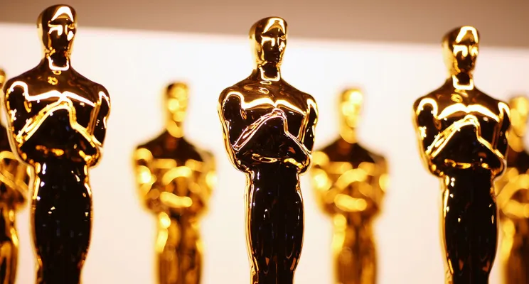 Close-up of multiple golden Oscar statuettes in a row with a white and beige background.