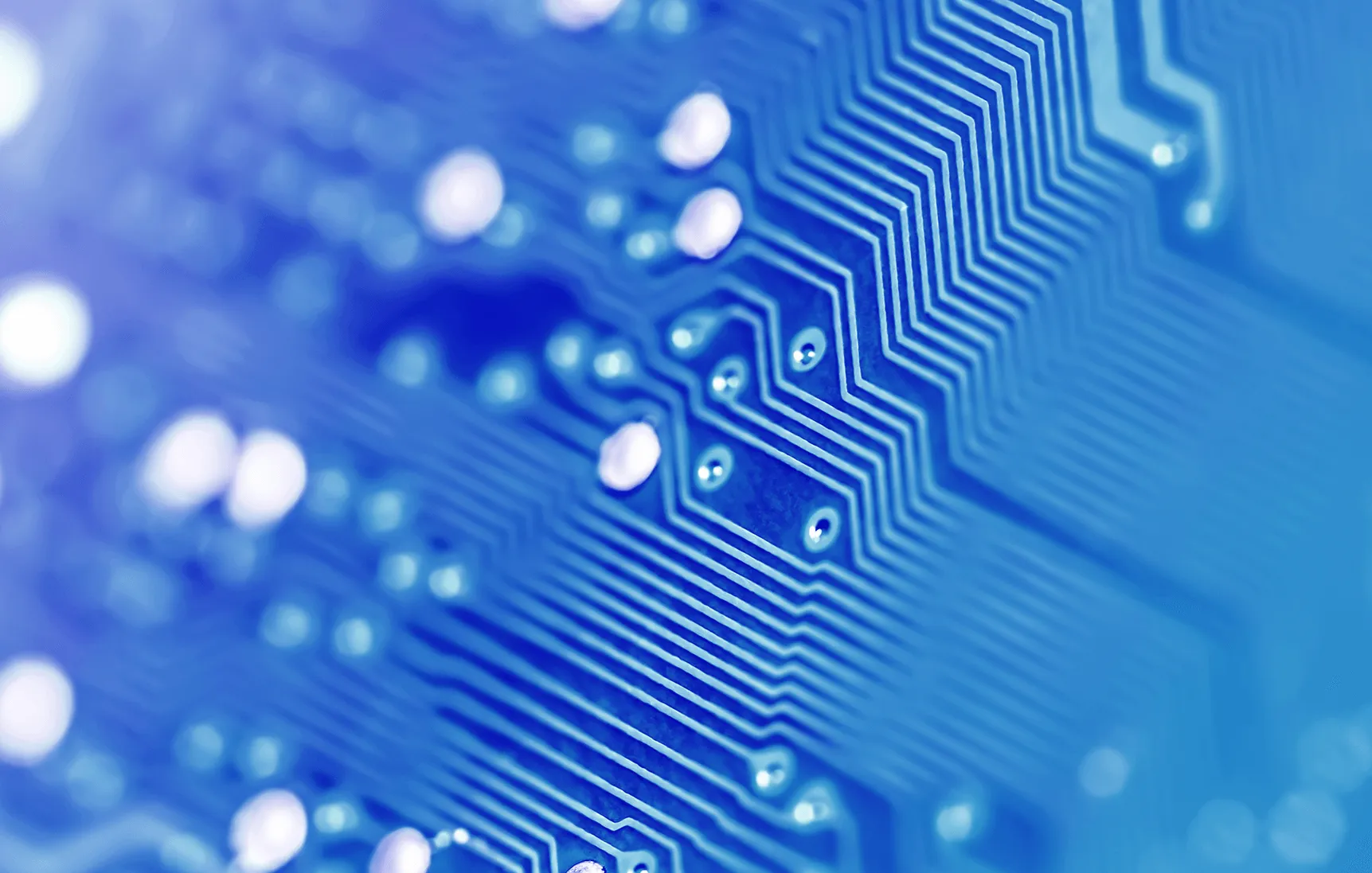 Close-up of a blue circuit board showing intricate white conductive pathways and solder points with a blurred background.