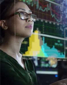A woman wearing glasses looks intently at multiple computer screens displaying colorful financial charts and data visualizations in a dimly lit room.