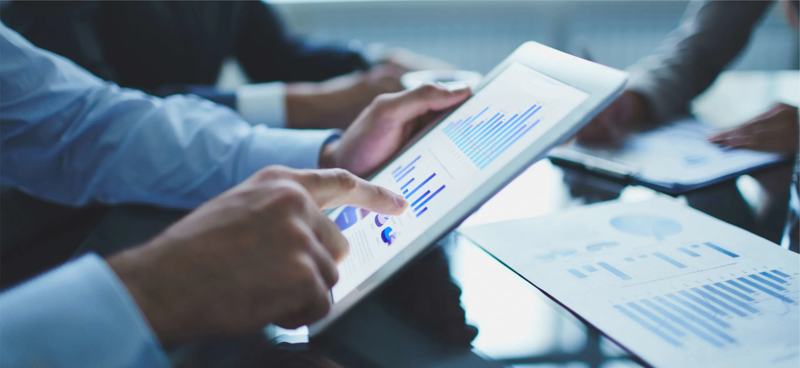 Close-up of a person using a tablet displaying blue and purple business charts and graphs, with printed charts and another person holding documents in the background.