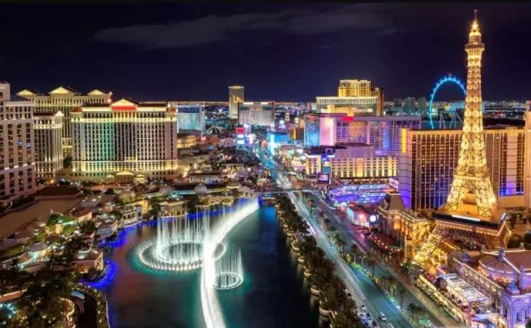 Night view of the Las Vegas Strip with illuminated buildings, a fountain show in a lake, and a replica of the Eiffel Tower on the right side.