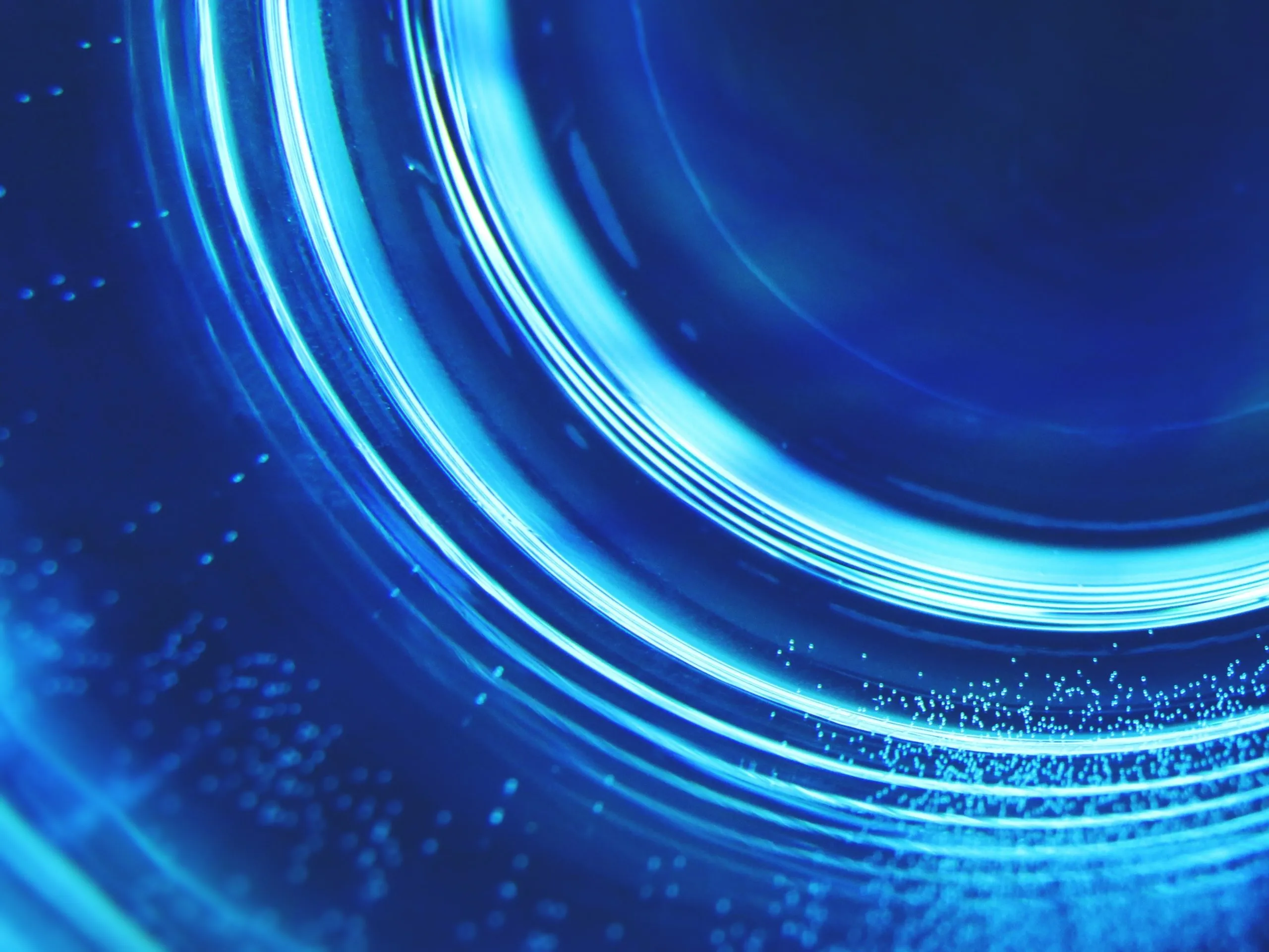 Close-up of concentric bright blue light rings and small illuminated dots on a dark blue background, resembling the inside of a glass or a lens reflecting light.