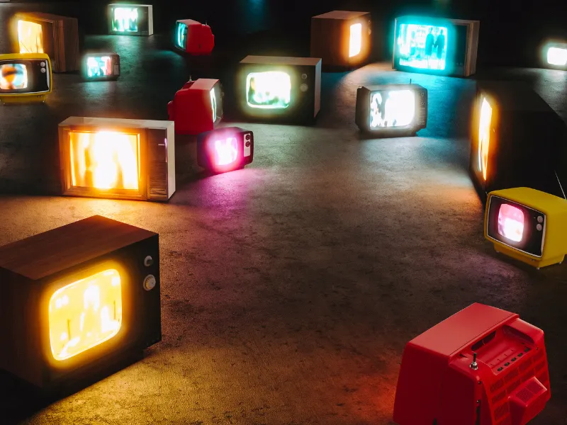 A dark room with multiple vintage televisions of various sizes and colors scattered on the floor, all displaying bright, blurry images emitting warm and cool glows.