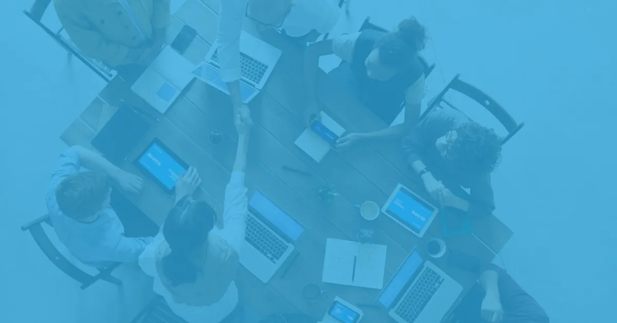 Overhead view of six people sitting around a wooden table using laptops, tablets, and smartphones, with two individuals shaking hands in the center, indicating a collaborative business meeting.