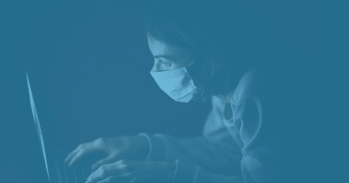 Person wearing a face mask and working on a laptop with focused expression.