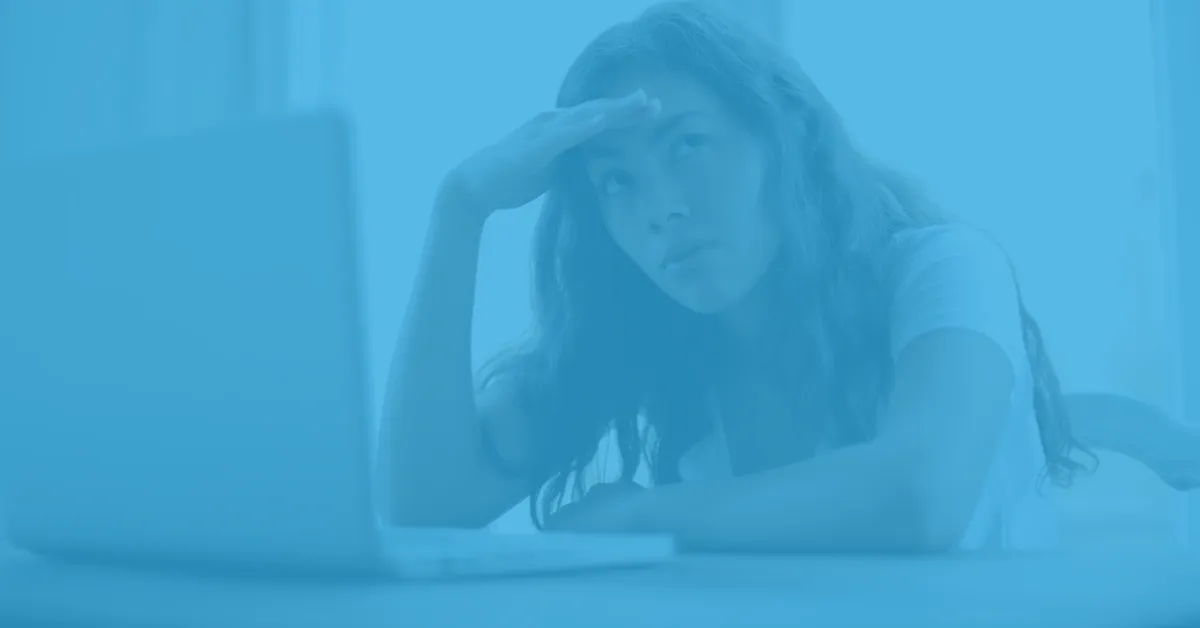 Young woman with long hair sitting at a table in front of a laptop, resting her head on her arm and shielding her eyes with her hand while looking up thoughtfully.