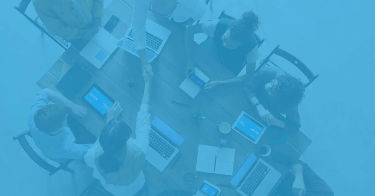 Overhead view of a group of people sitting around a wooden table with laptops, tablets, and notebooks, two individuals shaking hands over the table.